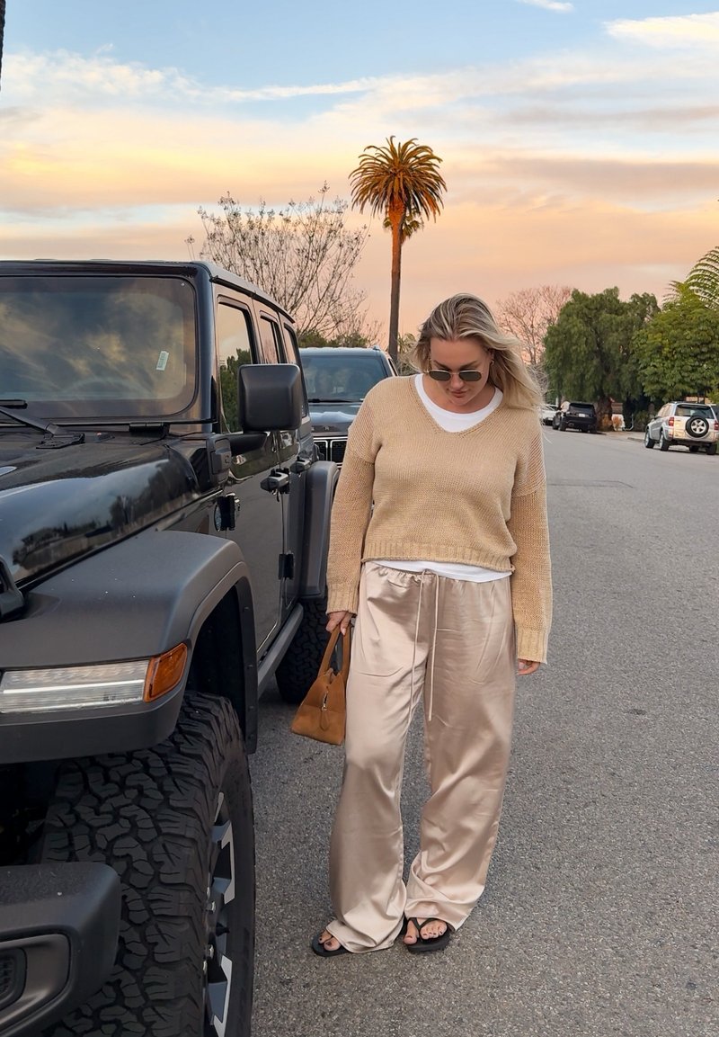 Woman in beige jumper and satin trousers with sunglasses holds a brown bag standing on street near black SUV at sunset.