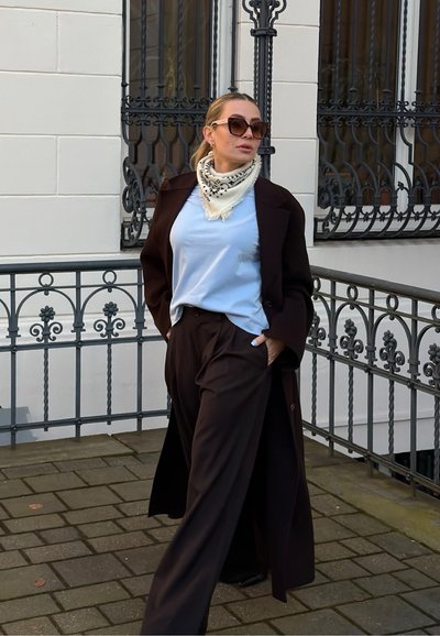Mujer con gafas de sol, camisa azul claro, pañuelo blanco con estampado, abrigo oscuro y pantalones, de pie en una zona pavimentada junto a barandillas decorativas de hierro.