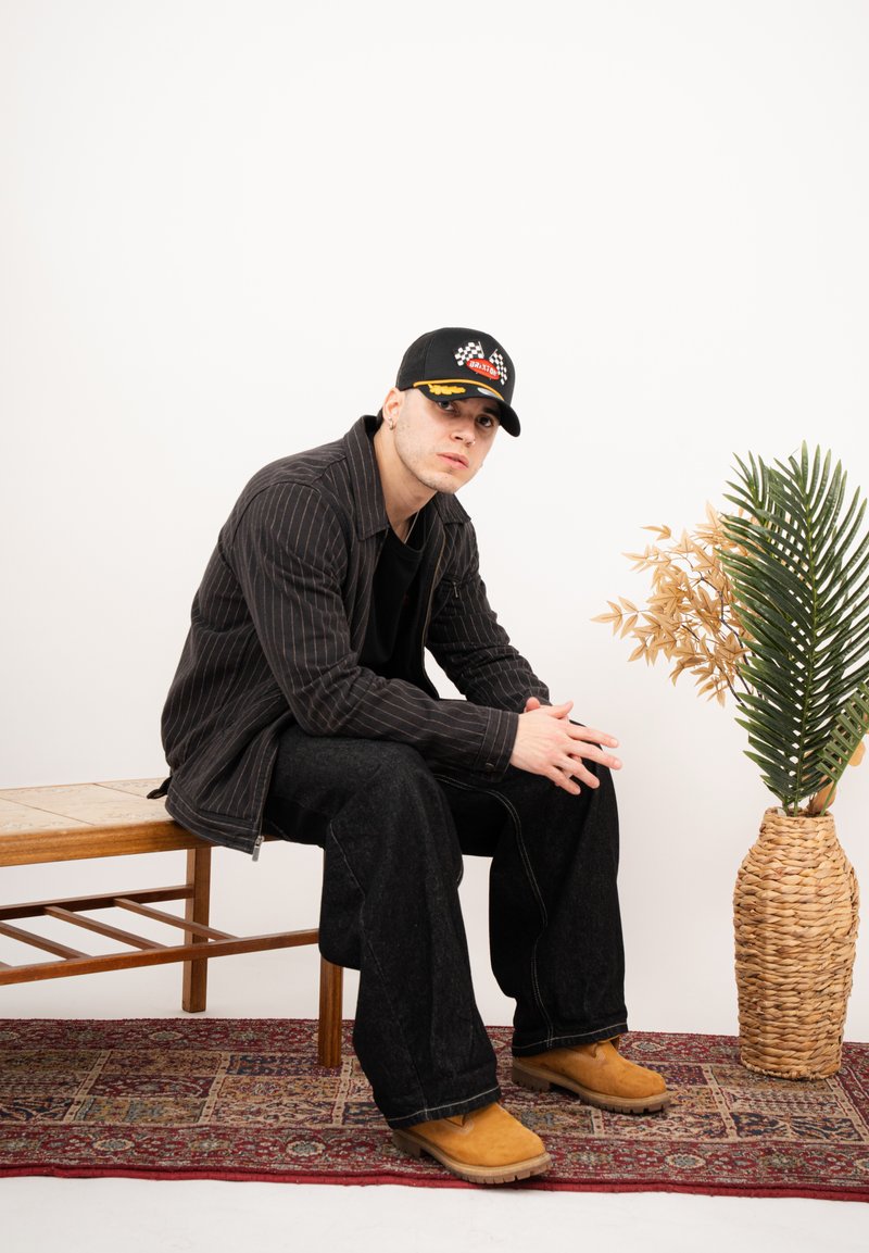 Tânăr purtând o șapcă neagră, jachetă cu dungi, pantaloni negri și bocanci căprui, stând pe o bancă de lemn lângă un coș împletit cu plante verzi și uscate.