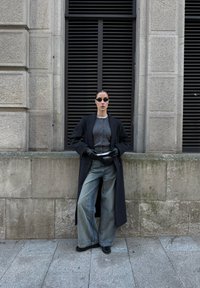 Femme portant des lunettes de soleil noires, un long manteau noir, un pull gris, un jean à jambes larges et des gants noirs, debout devant un bâtiment en pierre avec des grilles verticales.