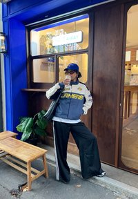 Veste Lakers bleue et blanche, pantalon large noir avec des bandes blanches, casquette noire, tenant une tasse de café, debout près d'un banc en bois et de plantes.