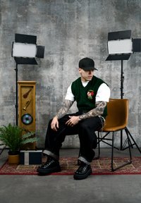 Jeune homme tatoué portant une casquette noire et un gilet vert assis sur une chaise marron entre des éclairages de studio et une décoration musicale sur un tapis.