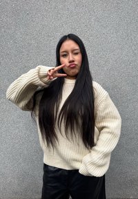 Pullover oversize in crema a maglia con polsini a coste, abbinato a pantaloni neri, posato davanti a un muro testurizzato grigio. Gesto della pace con la mano destra.