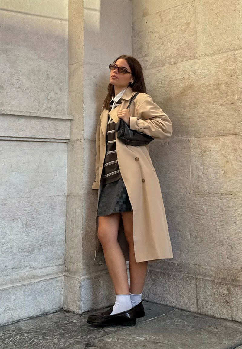 Nainen, jolla on päällään beige trench coat, raidallinen paita, lyhyt hame, valkoiset sukat ja mustat kengät, seisoo kiviseinää vasten pitäen pientä mustaa laukkua.