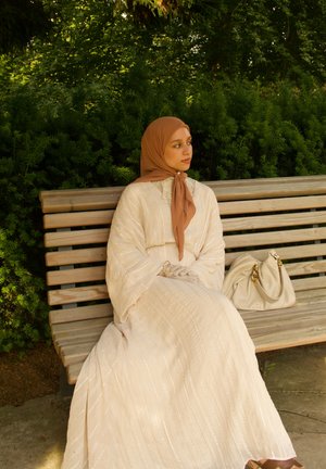 Ilga, kreminės spalvos suknelė su subtiliomis vertikaliomis juostelėmis ir plačiomis rankovėmis. Smėlio spalvos hidžabas ir biski nagrinėjantis rankinukas puikiai papildydami išvaizdą, sėdint ant medinės suolo.