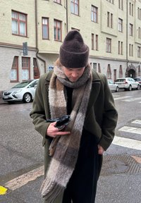 Homme portant un manteau vert, un bonnet en tricot marron et une grande écharpe à carreaux tient des gants noirs en marchant dans une rue de la ville mouillée près de voitures garées.