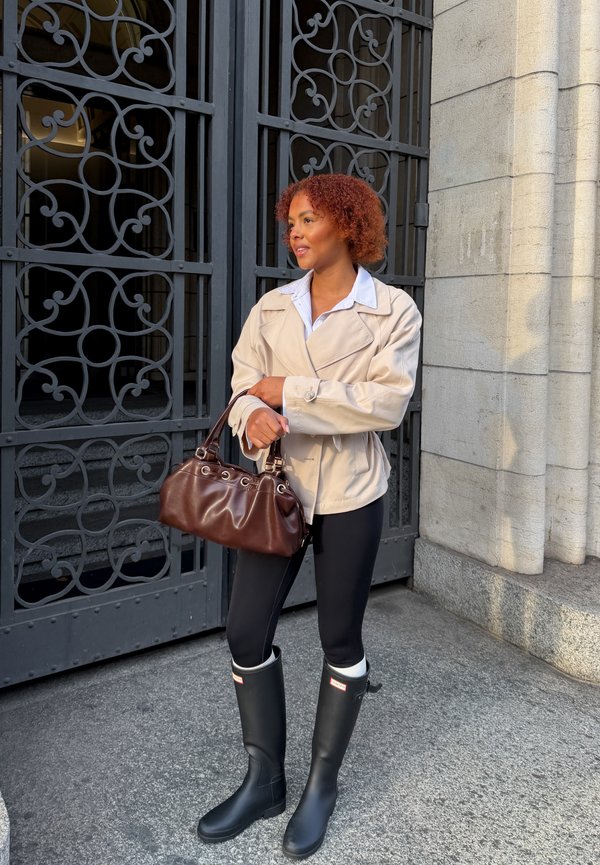 Donna con giacca beige, leggings neri e stivali da pioggia neri che tiene una borsa marrone, in piedi vicino a un cancello in metallo nero ornato e a un muro di pietra.