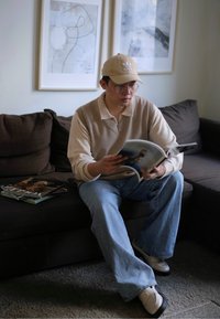 Beige knitted jumper, light blue wide-leg jeans, silver loafers, clear glasses, and a beige cap. Reading a magazine on a dark sofa.