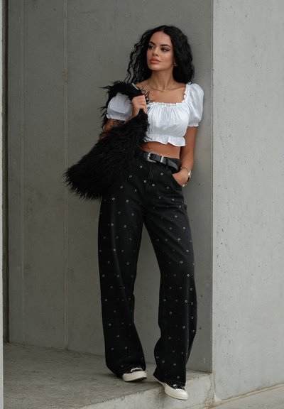 Jeans negros con detalles de ojales metálicos, de corte holgado, combinados con una blusa blanca corta. Los accesorios incluyen un bolso negro texturizado.