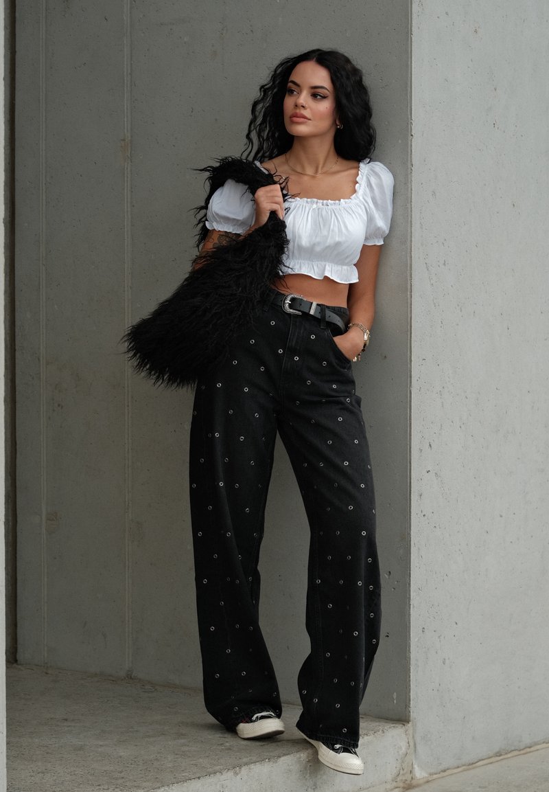 Schwarze Jeans mit metallischen Ösenakzenten, lockerer Schnitt, kombiniert mit einer weißen cropped Bluse. Zubehör umfasst eine strukturierte schwarze Handtasche.