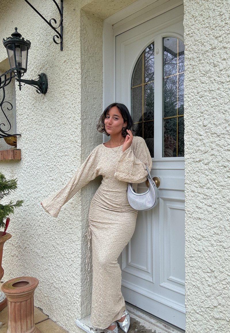 Young woman in beige textured long dress and silver shoes poses by white door, holding small silver handbag with outdoor lantern and plants nearby.