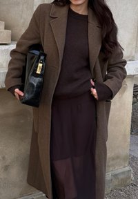 Manteau en laine marron avec une coupe ajustée, associé à un pull en maille marron et un sac en cuir noir orné de détails dorés.