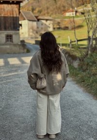 Nadmerný sivý vlnený kabát, krémové široké nohavice a strieborná textúrovaná crossbody taška. Dlhé hnedé vlasy a štrkova cesta v malebnom prostredí.