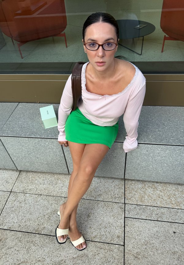 Femme avec des lunettes assise sur un banc en pierre à l'extérieur, portant un haut à manches longues rose pâle, une jupe verte et des sandales crème à enfiler.