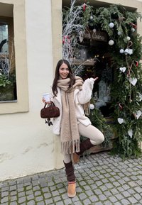 Jeune femme en vêtements d'hiver tenant une tasse de café et un sac à main marron, posant joyeusement à côté d'une vitrine de Noël décorée.