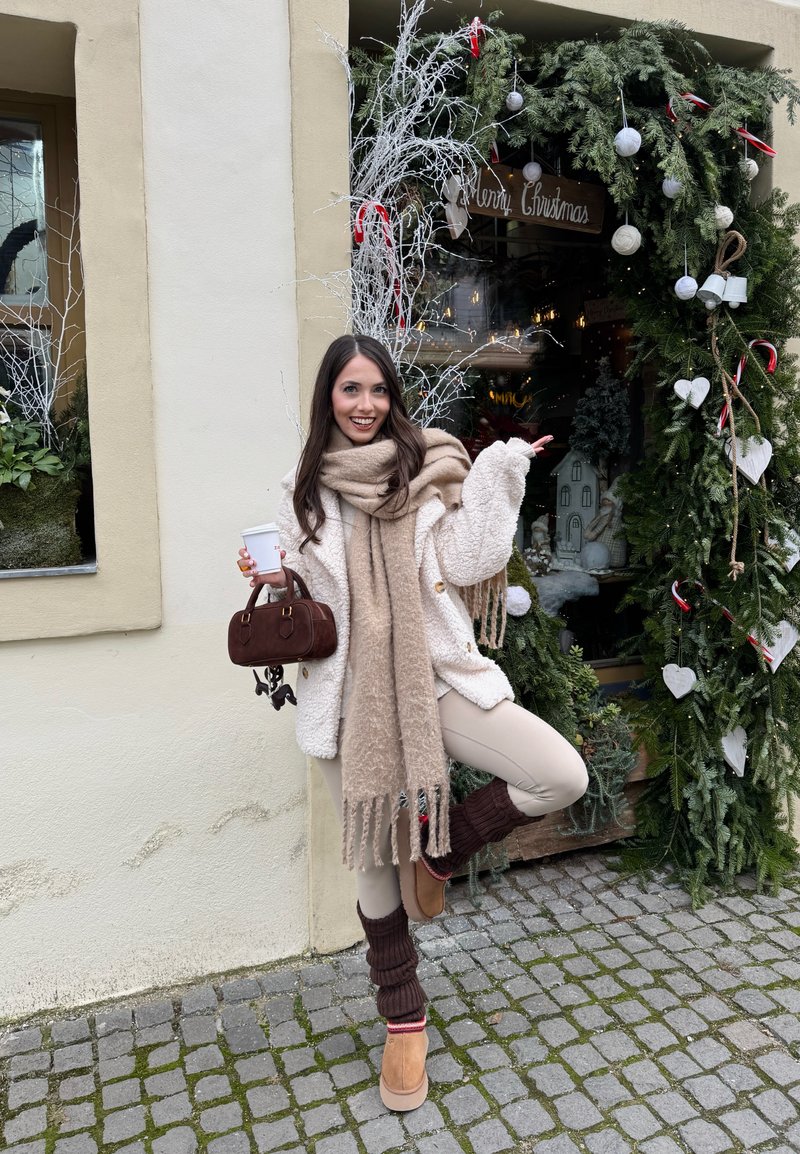 Jeune femme en vêtements d'hiver tenant une tasse de café et un sac à main marron, posant joyeusement à côté d'une vitrine de Noël décorée.