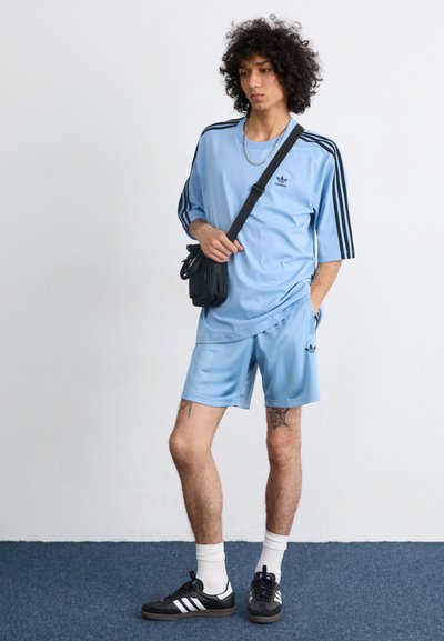 Conjunto deportivo azul claro con acentos negros; camiseta de manga corta holgada y pantalones cortos. Presenta un bolso cruzado negro y zapatillas negras.