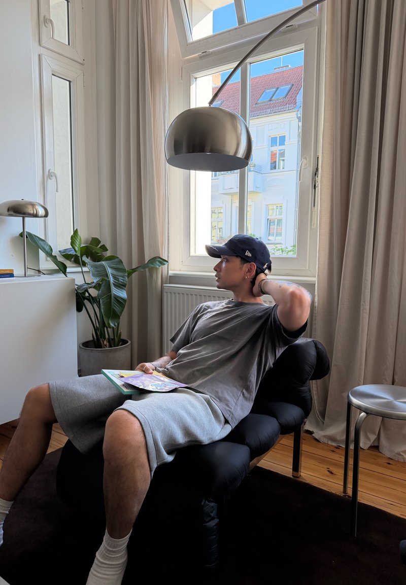 Jeune homme en casquette se détend sur une chaise noire tenant un livret, à côté d'une grande fenêtre avec des rideaux beige, une plante et une lampe sur pied moderne dans une pièce lumineuse.