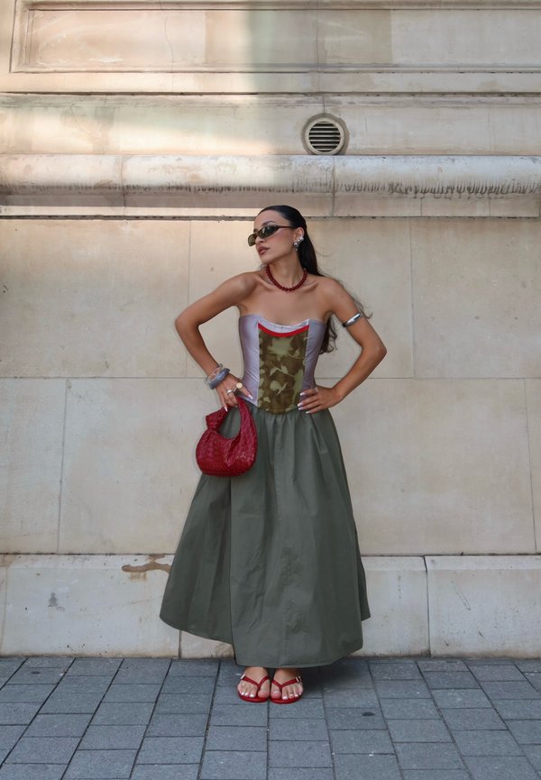 Femme en robe verte et grise sans bretelles avec des accents rouges, tenant un sac à main rouge, portant des lunettes de soleil et des sandales rouges, debout contre un mur beige.