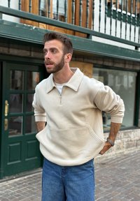 Beige hettegenser med glidelås, stor krage og frontlomme, kombinert med blå denimjeans og klokke. Avslappet, myk stofftekstur.