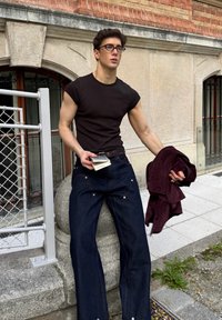 Czarna dopasowana koszulka, ciemnoniebieskie jeansy z szerokimi nogawkami i kontrastowym przeszyciem, trzymająca bordo kurtkę i mały notes, w plenerze.