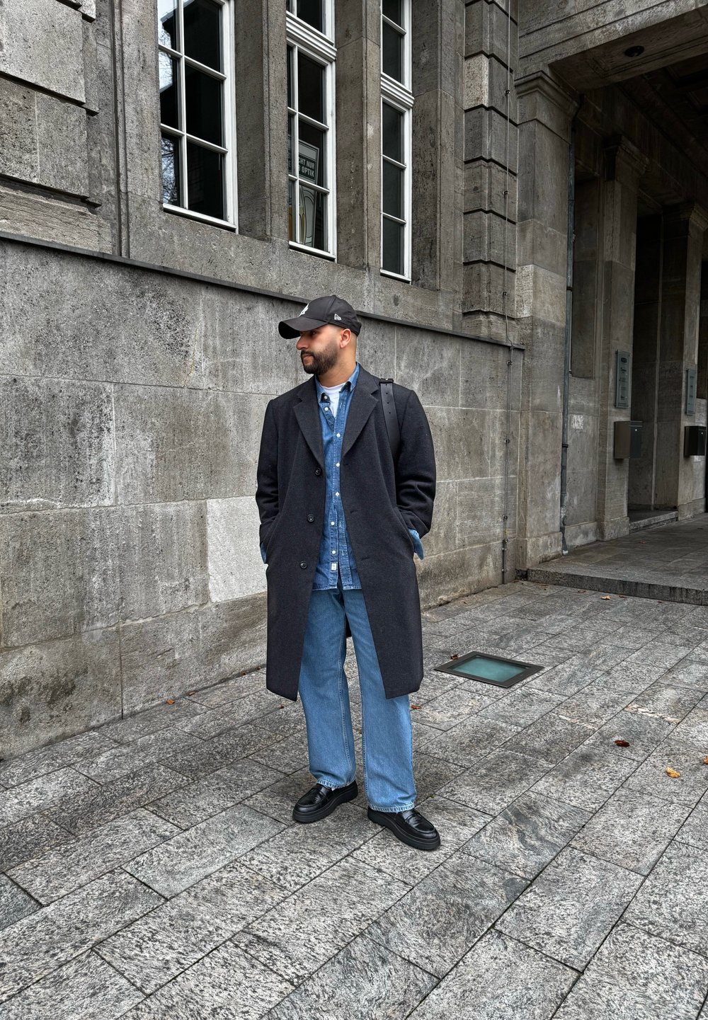 Σκούρο γκρι μάλλινο παλτό, μπλε τζιν πουκάμισο και φαρδιά τζιν παντελόνια. Μαύρο καπέλο μπέιζμπολ και παπούτσια. Σταθείτε σε ανάγλυφη πέτρινη επιφάνεια.