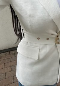 Blazer blanco texturizado con un bolsillo lateral, que presenta un cinturón con herrajes dorados y una silueta ajustada. Adecuado para diversas ocasiones.