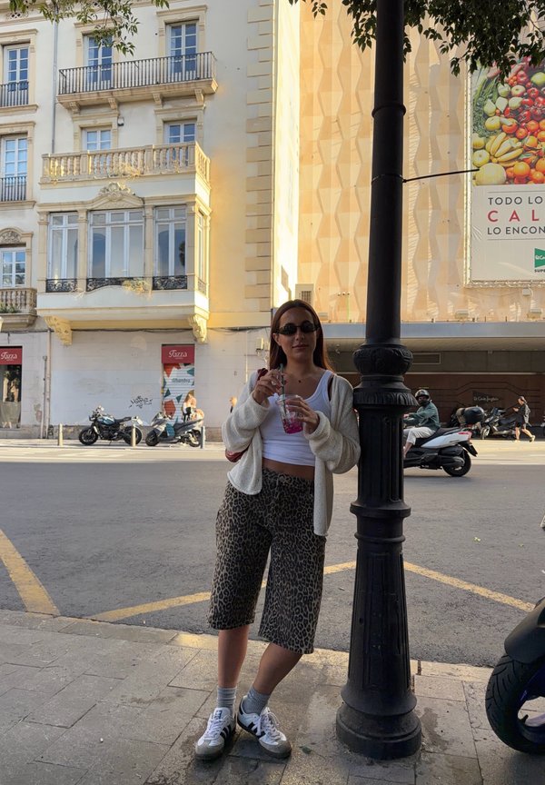 Femme portant des lunettes de soleil, un haut blanc et un short à imprimé léopard, buvant dans une tasse, appuyée contre un lampadaire noir sur un trottoir en ville.