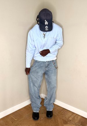 Persona que lleva una camisa abotonada azul claro, vaqueros sueltos azul claro con una cadena de cuentas, botas negras y una gorra de béisbol negra de LA mirando hacia abajo.