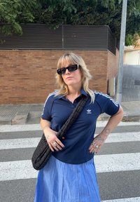 Camisa polo azul marinho com riscas brancas, mala preta à tiracolo e saia plissada azul clara; tatuagens visíveis nas mãos; óculos de sol usados.