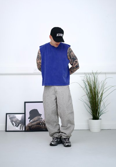 Top sin mangas azul en tejido suave, combinado con pantalones grises claros y texturizados. Una gorra negra y zapatillas completan el look. Entorno interior.