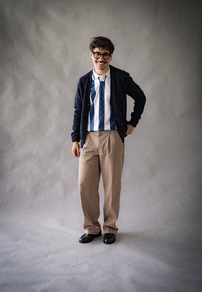 Cardigan navy sobre una camisa a rayas azul y blanca, pantalones beiges y zapatos negros. Hombre de pie con una mano en la cadera contra un fondo gris.