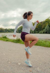 Žena u bijeloj dugoj majici i ljubičastim šortsevima vježba podižući jednu nogu na šljunčanom putu uz drveće i vodu pod oblačnim nebom.