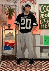 Črna NFL majica z belimi detajli, sivi trenirka in črne superge. V ozadju so police s čevlji, rastlinami in dekorativnimi predmeti.