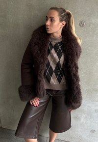 Veste avec fourrure brune sur un pull à motif argyle et culottes en cuir brun, présentant un design élégant et des matériaux texturés.