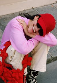 Pulover roz, căciulă roșie, ochelari de soare supradimensionați, pantaloni bej și o geantă accentuată cu flori roșii. Zâmbind cu brațele încrucișate, așezată în aer liber.