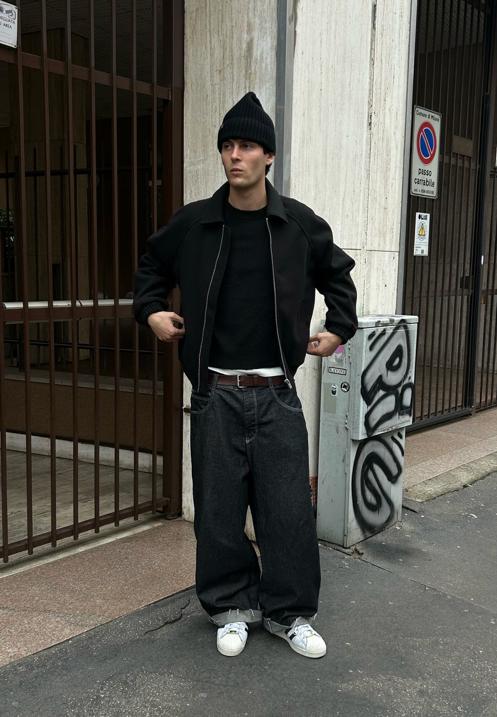 Jeune homme portant un bonnet noir, une veste noire, un jean large foncé et des baskets blanches, debout sur un trottoir près d'un bâtiment fermé par une grille et d'une boîte utilitaire couverte de graffitis.