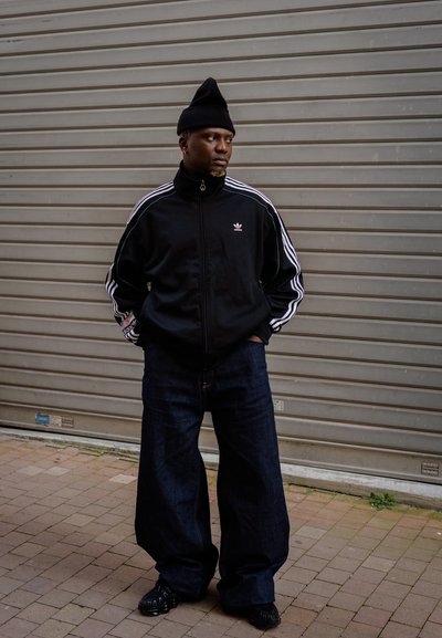 Chaqueta de chándal negra con cierre de cremallera y rayas blancas, combinada con vaqueros de denim azules sueltos. Un gorro negro y zapatos oscuros completan el atuendo.