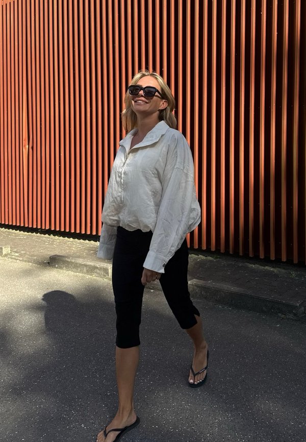 Frau mit Sonnenbrille, weißem Langarmshirt, schwarzen kurzen Hosen und Flip-Flops, die auf dem Gehsteig neben einer orangefarbenen vertikal lamellierten Wand geht.