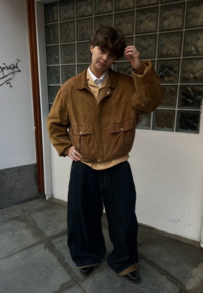 Persona con cabello corto y castaño se acomoda el cabello, lleva una chaqueta marrón, camisa de cuadros, pantalones anchos negros y zapatos negros, de pie junto a una pared de bloques de vidrio.