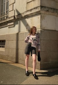 Sweter w różowo-szary kratkę, czarne dżinsowe szorty i czarne loafersy. Stoi przy teksturowanej kamiennej ścianie, słońce rzuca cienie.