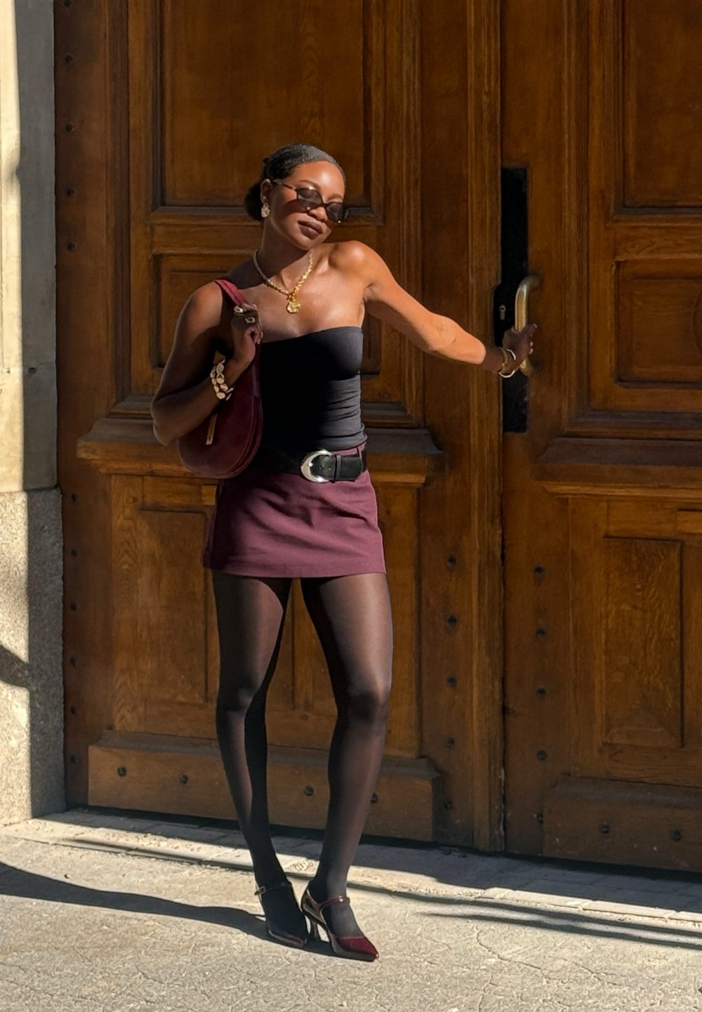 Nais, kes kannab musta ilma õlata topi, burgundi seelikut ja musti sukki, hoides burgundi kotti. Tal on prillid ja kuldne ehe. Puituks taustal.