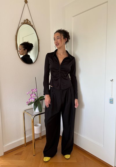 Mujer con cabello rizado que lleva una blusa negra fruncida y pantalones anchos negros, de pie junto a una pequeña mesa con una orquídea rosa y un espejo ovalado en la pared.