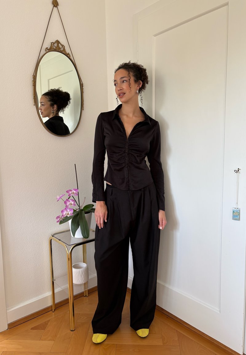 Femme aux cheveux bouclés portant un chemisier noir froncé et un pantalon large noir, debout près d'une petite table avec une orchidée rose et un miroir ovale accroché au mur.