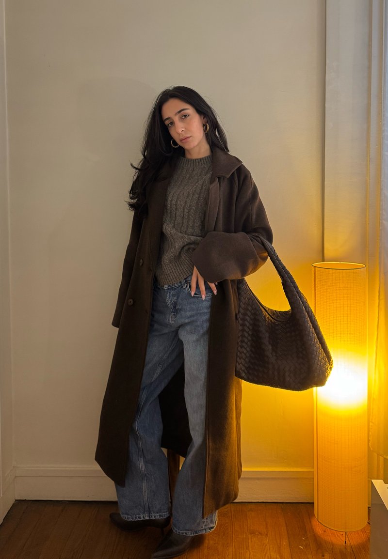 Vrouw met een lange donkere jas, grijze trui, brede jeans en zwarte laarzen, die een grote geweven handtas vasthoudt, staand naast een hoge verlichte vloerlamp.