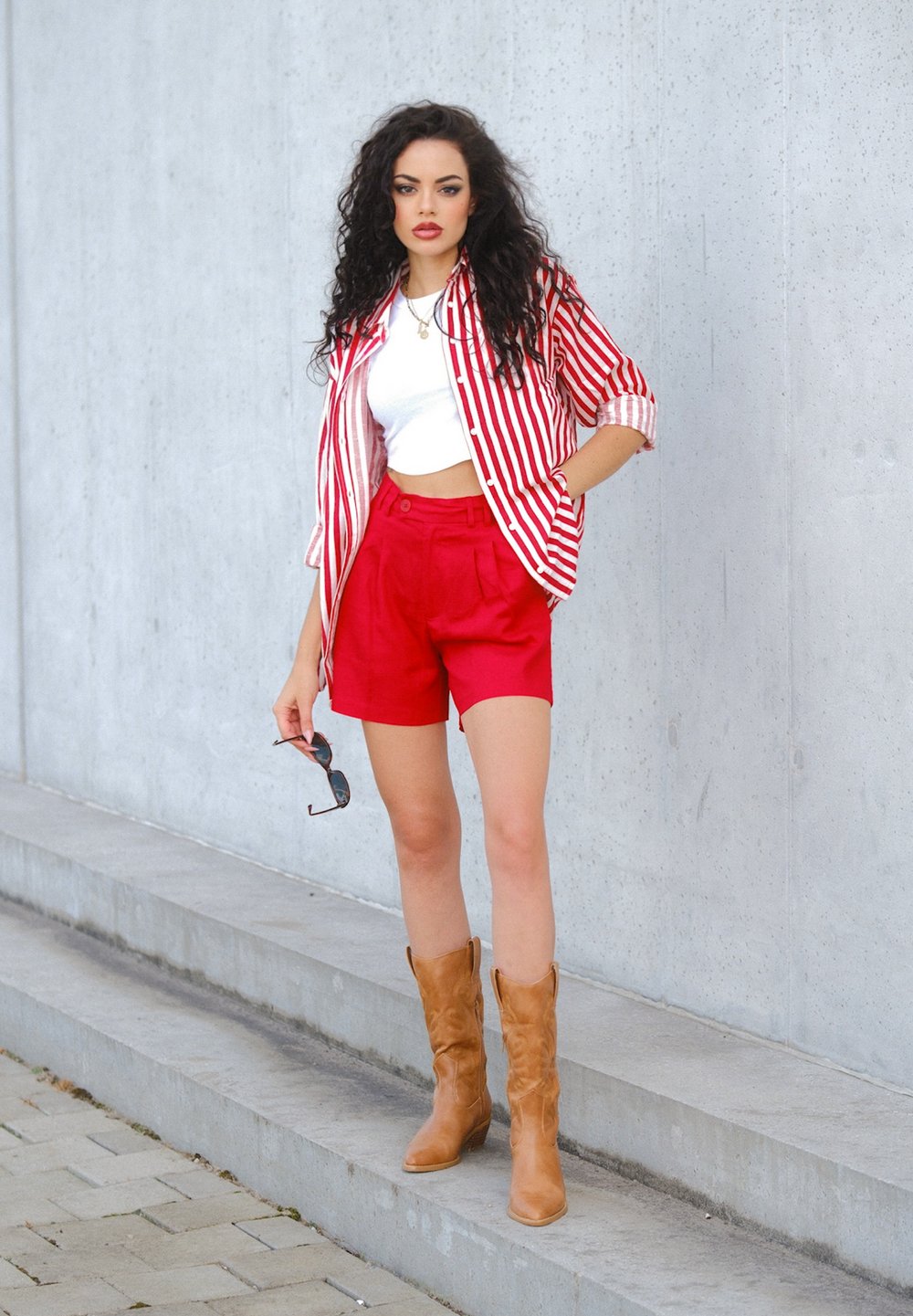 Maglietta a righe rosse e bianche, top corto bianco, pantaloni corti rossi e stivali da cowboy color sabbia. Regge degli occhiali da sole. Sfondo grigio uniforme con gradini in cemento.