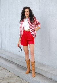 Maglietta a righe rosse e bianche, top corto bianco, pantaloni corti rossi e stivali da cowboy color sabbia. Regge degli occhiali da sole. Sfondo grigio uniforme con gradini in cemento.