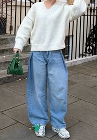 Pull blanc en maille à col en V, jean large en denim bleu décontracté, sac à main vert et baskets argentées avec des accents à motifs.