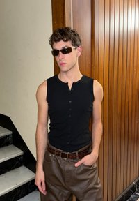 Young man wearing rectangular sunglasses, sleeveless black buttoned top, and brown leather pants, standing indoors by marble stairs and wooden panel wall.
