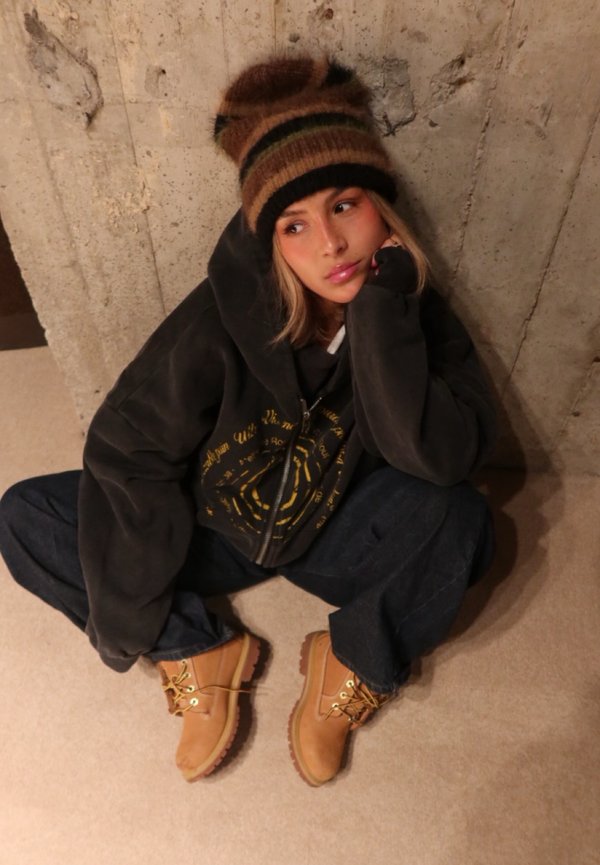 Sudadera gris oscuro de gran tamaño con gráfico amarillo, vaqueros azules sueltos, botas marrones con cordones y un gorro de rayas marrón y negro.